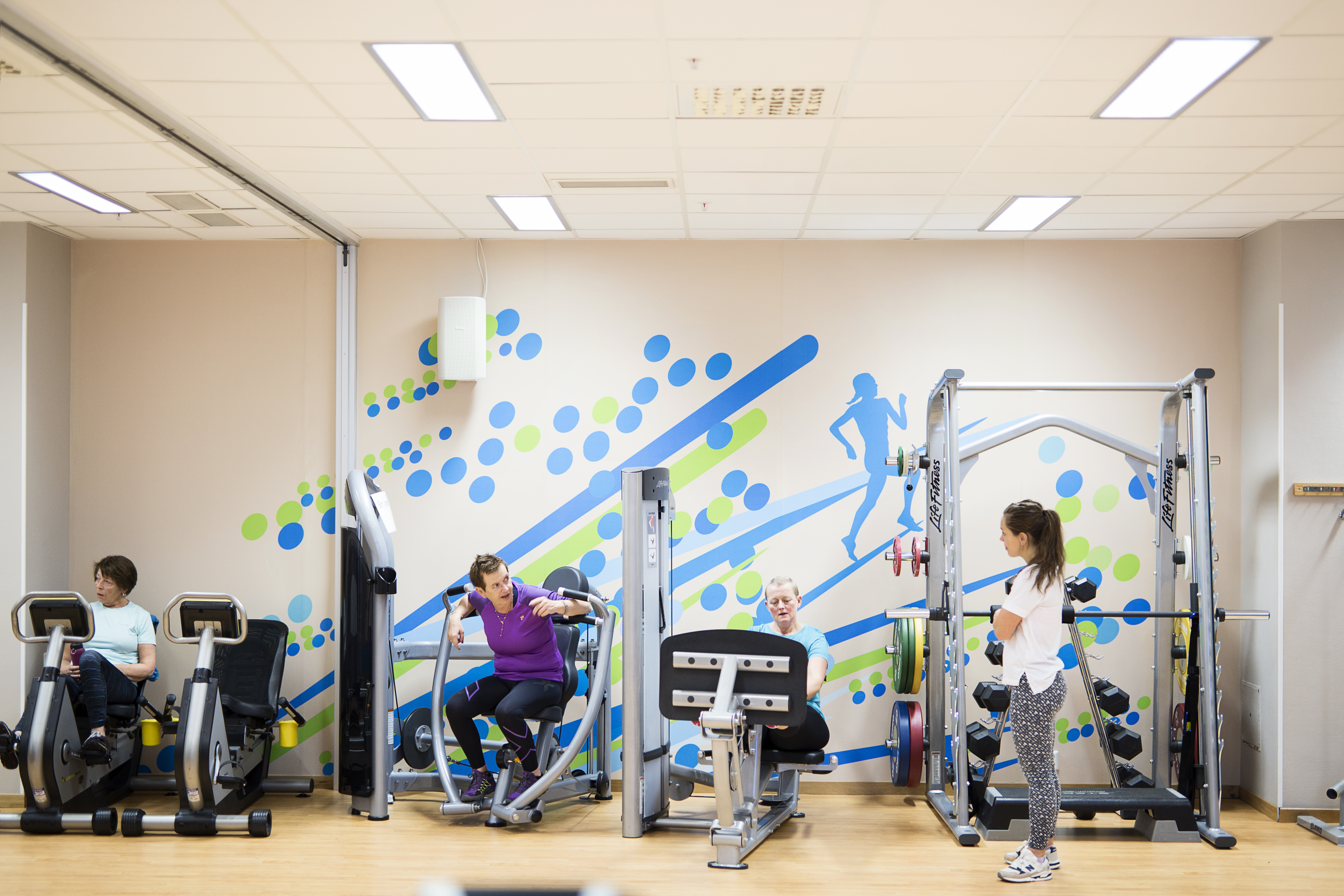 Tre kvinner trener på ulike treningsapparat med veiledning fra kvinnelig helse- og treningsfysiolog i treningsrom. Foto