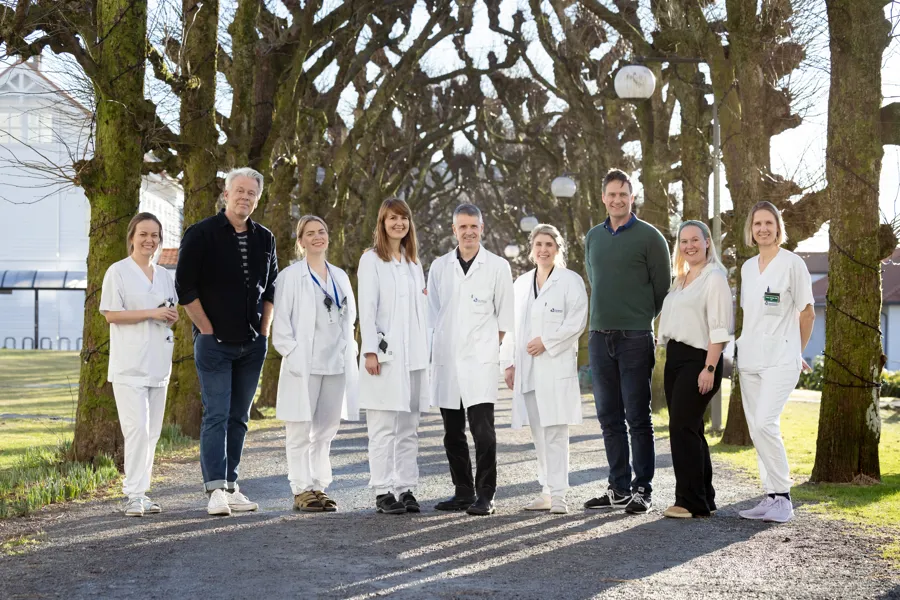 Gruppebilete med ni personar i legefrakkar som står ute i ein allé med tre.