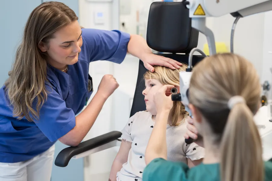 En person som undersøker øret til et barn, mens en annen voksen med uniform holder barnet.