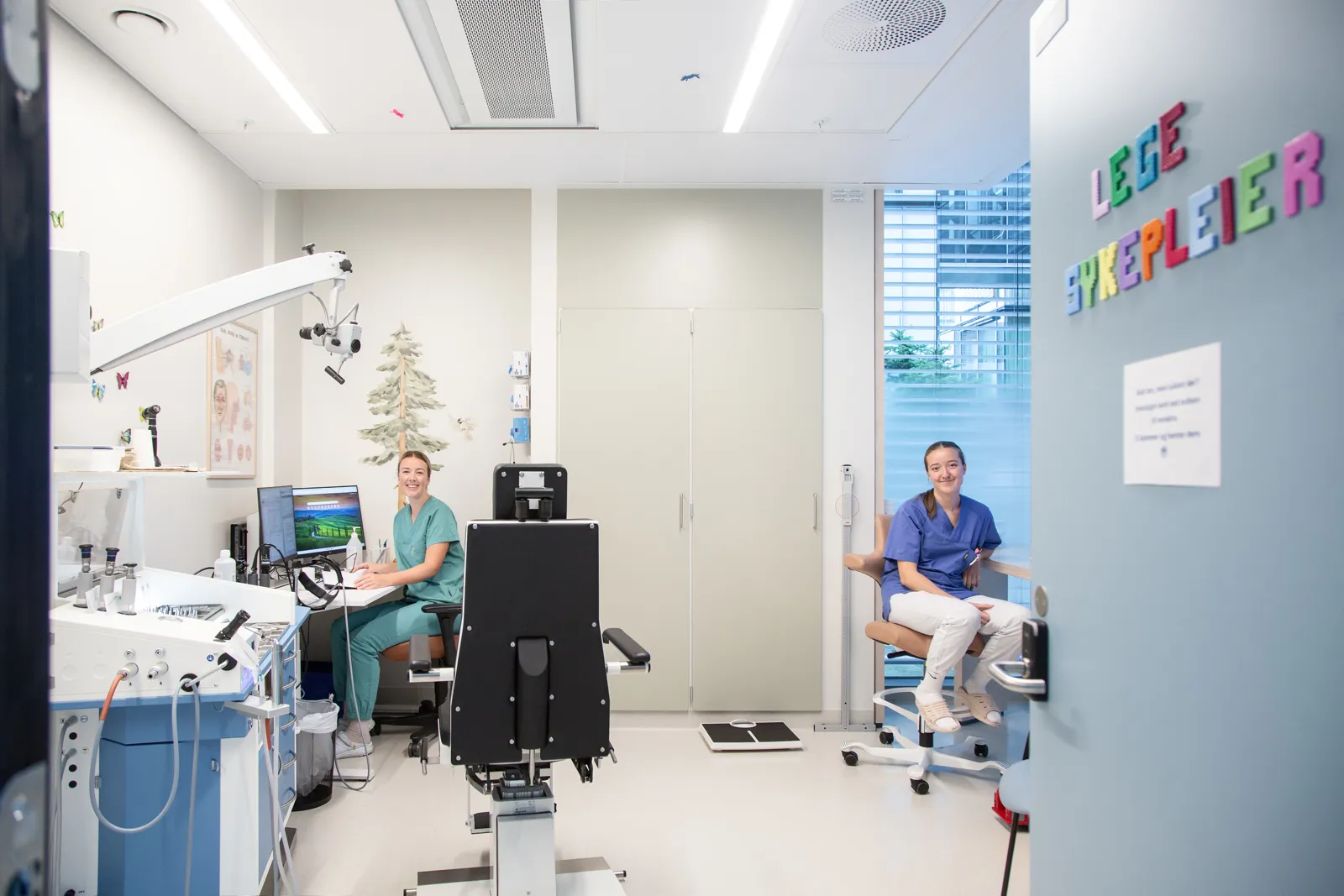 En lege og sykepleier i et rom med undersøkelsesstol, pc og medisinske apparater