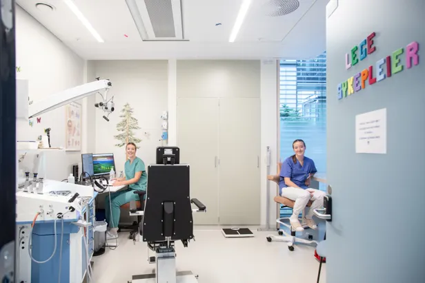 En lege og sykepleier i et rom med undersøkelsesstol, pc og medisinske apparater