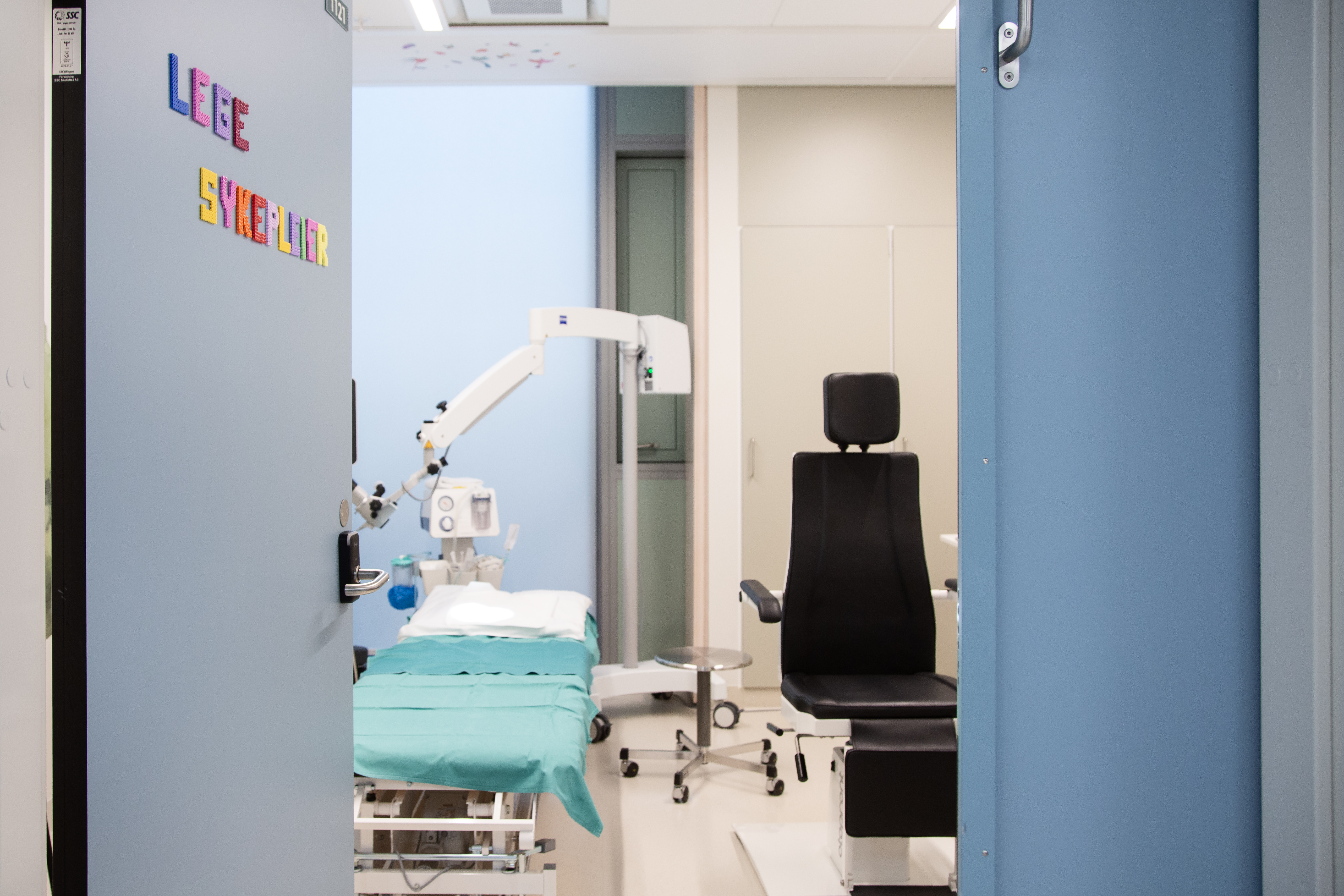 En åpen dør, der står skrevet Lege og sykepleier på, inn til et rom med en undersøkelsesbenk og -stol.