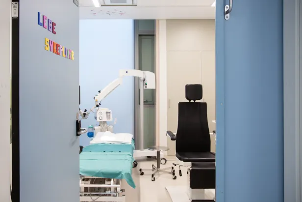 En åpen dør, der står skrevet Lege og sykepleier på, inn til et rom med en undersøkelsesbenk og -stol.