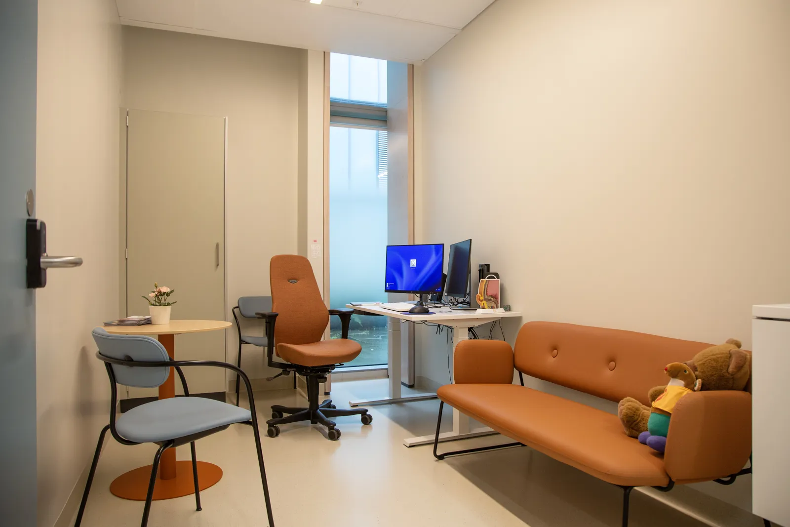 Et rom på sykehuset med flere stoler, en pc og en oransje sofa med en bamse i