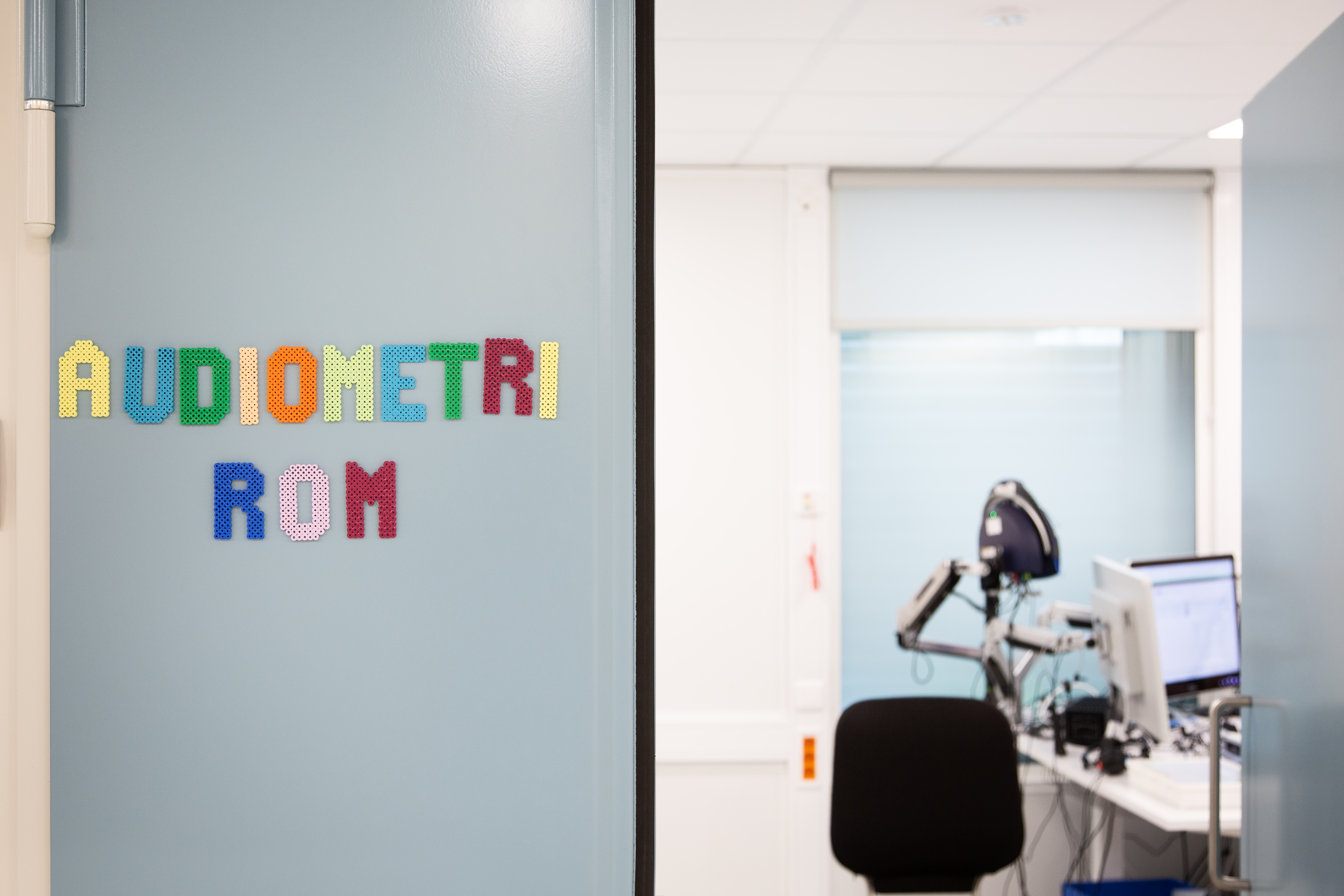 En åpen dør til et rom med stol, pc og medisinske apparater. Det står Audiometrirom i fargerik skrift på døren.