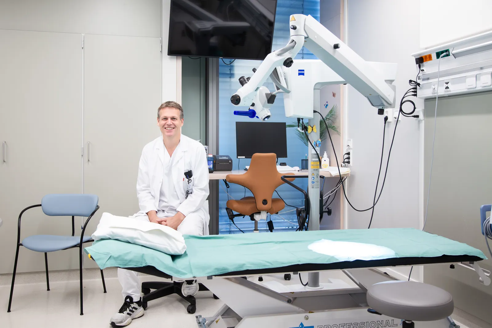 En mannlig lege som sitter smilende bak en undersøkelsesbenk.