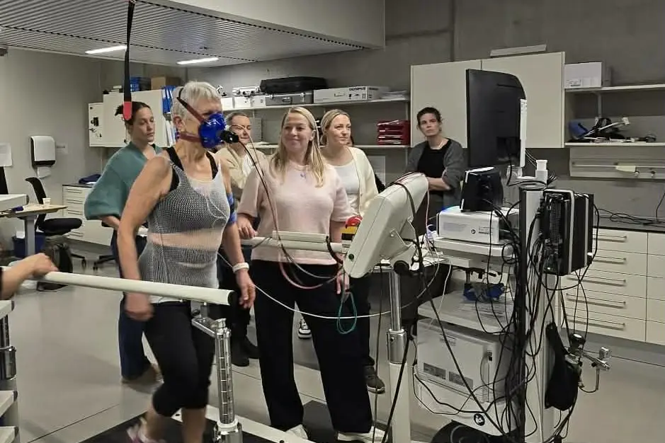 En kvinne som beveger seg på en tredemølle og har på en maske mens det står mennesker rundt