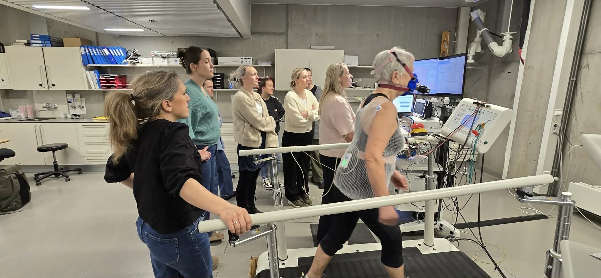 En kvinne med en maske og som beveger seg på en tredemølle mens det står flere personer bak henne
