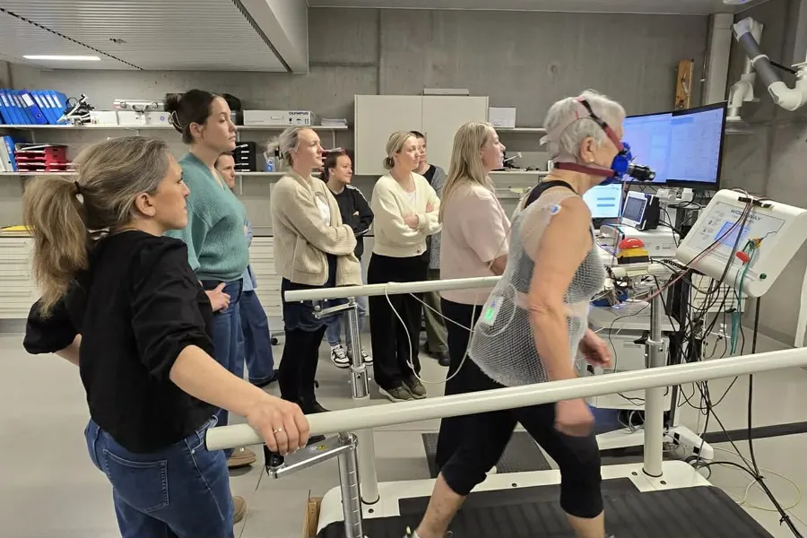 En kvinne med en maske og som beveger seg på en tredemølle mens det står flere personer bak henne