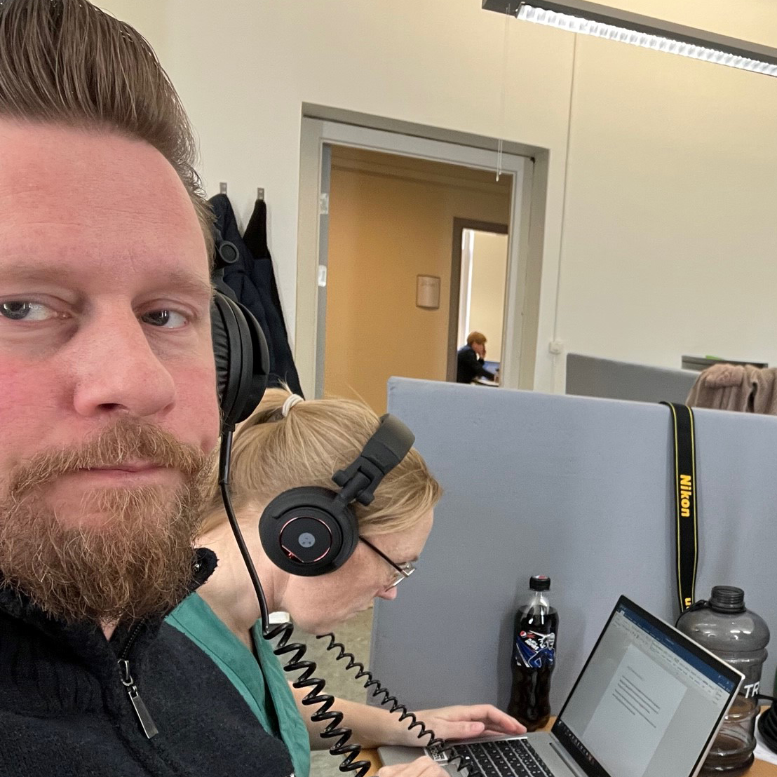 Portrett av en mann med hodetelefoner. I bakgrunnen ser du en kvinne som bøyer seg fram foran en PC