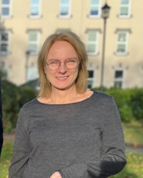 En kvinne med briller står fremfor bygg i en park.