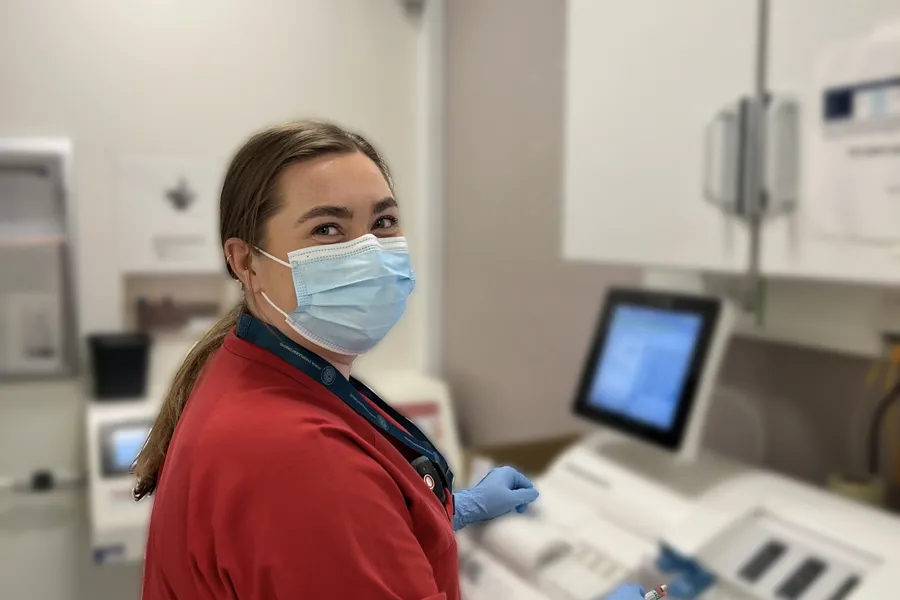 A woman wearing a mask and a red shirt in a room with white cabinets and a computer