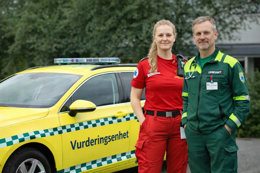 En mann i grønn uniform og en kvinne i rød uniform står ved siden av en gul bil det står vurderingsenhet på