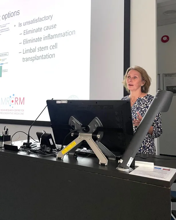 A person standing behind a desk with a laptop and a projector screen