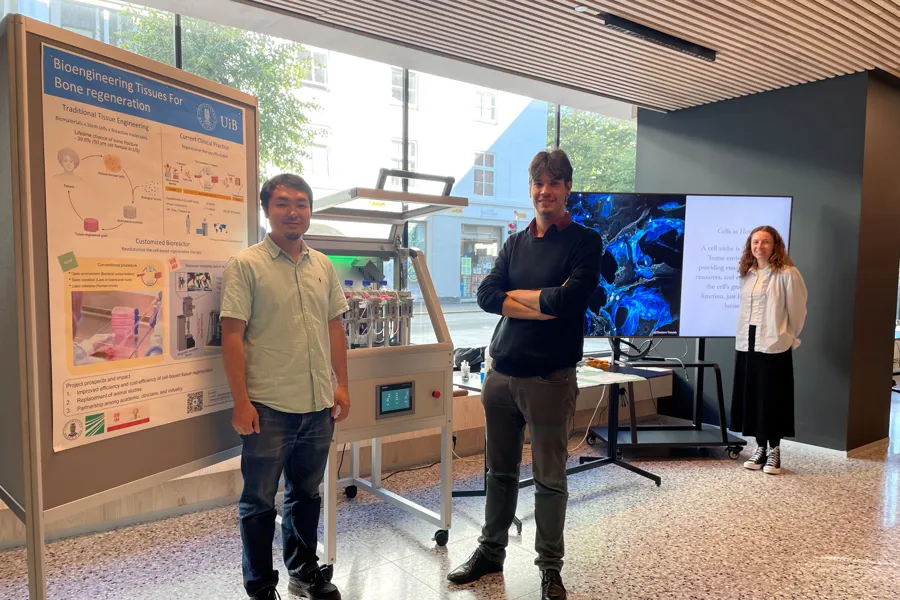 Two men and a woman standing next to a large poster, bioractor and photos on a screen.