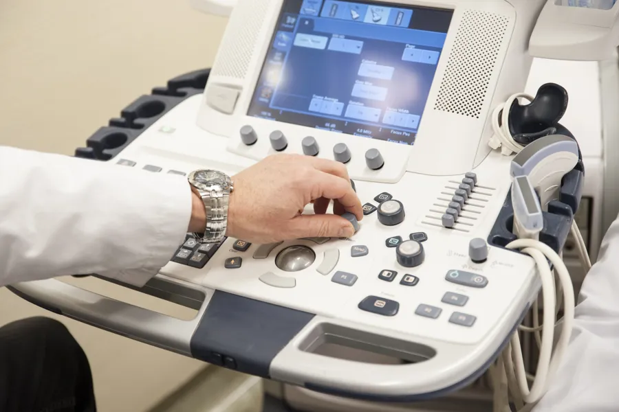 Hvitkledd arm med klokke rundt håndleddet og hånd som skrur på knott på panelet til ultralydmaskin. Foto