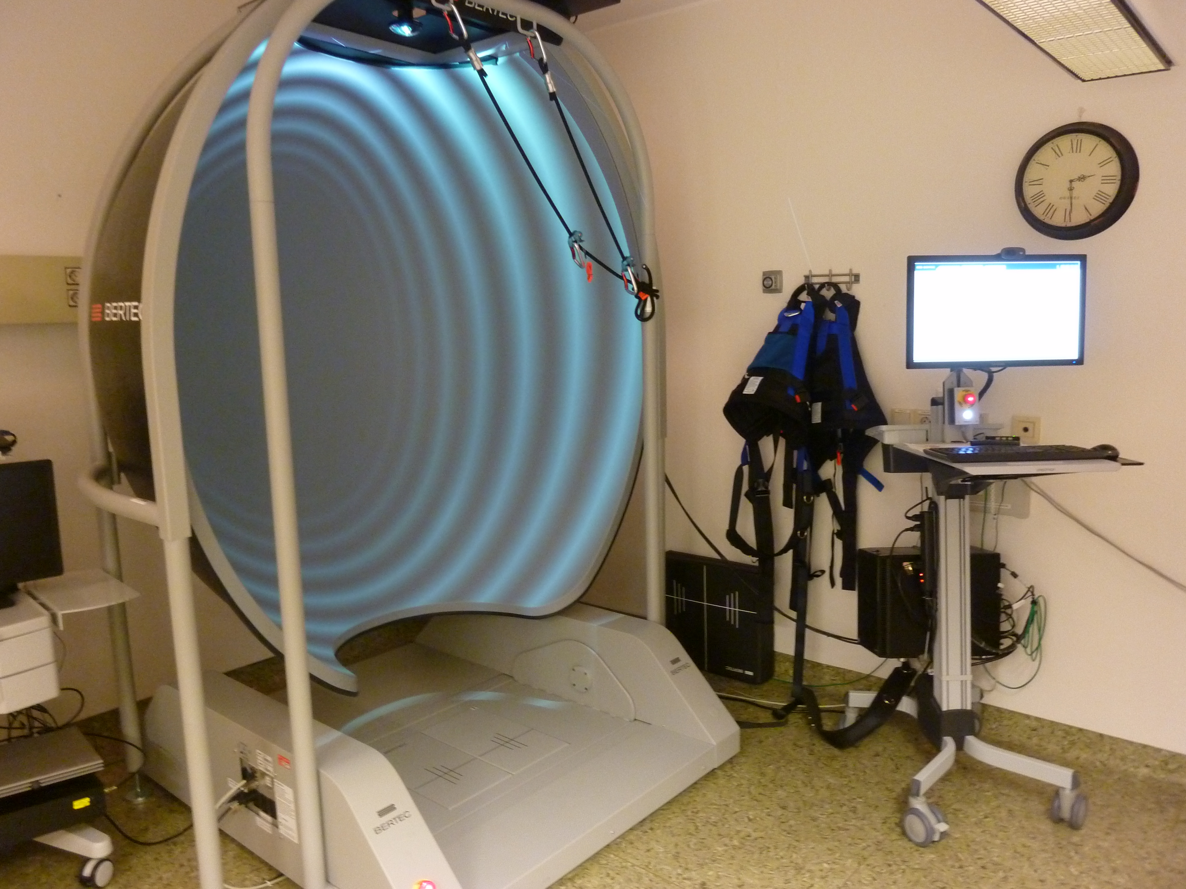 Interiør unndersøkelsesrom med balanseplattform og bord med PC-skjerm og tastatur, klokke på vegg. Foto