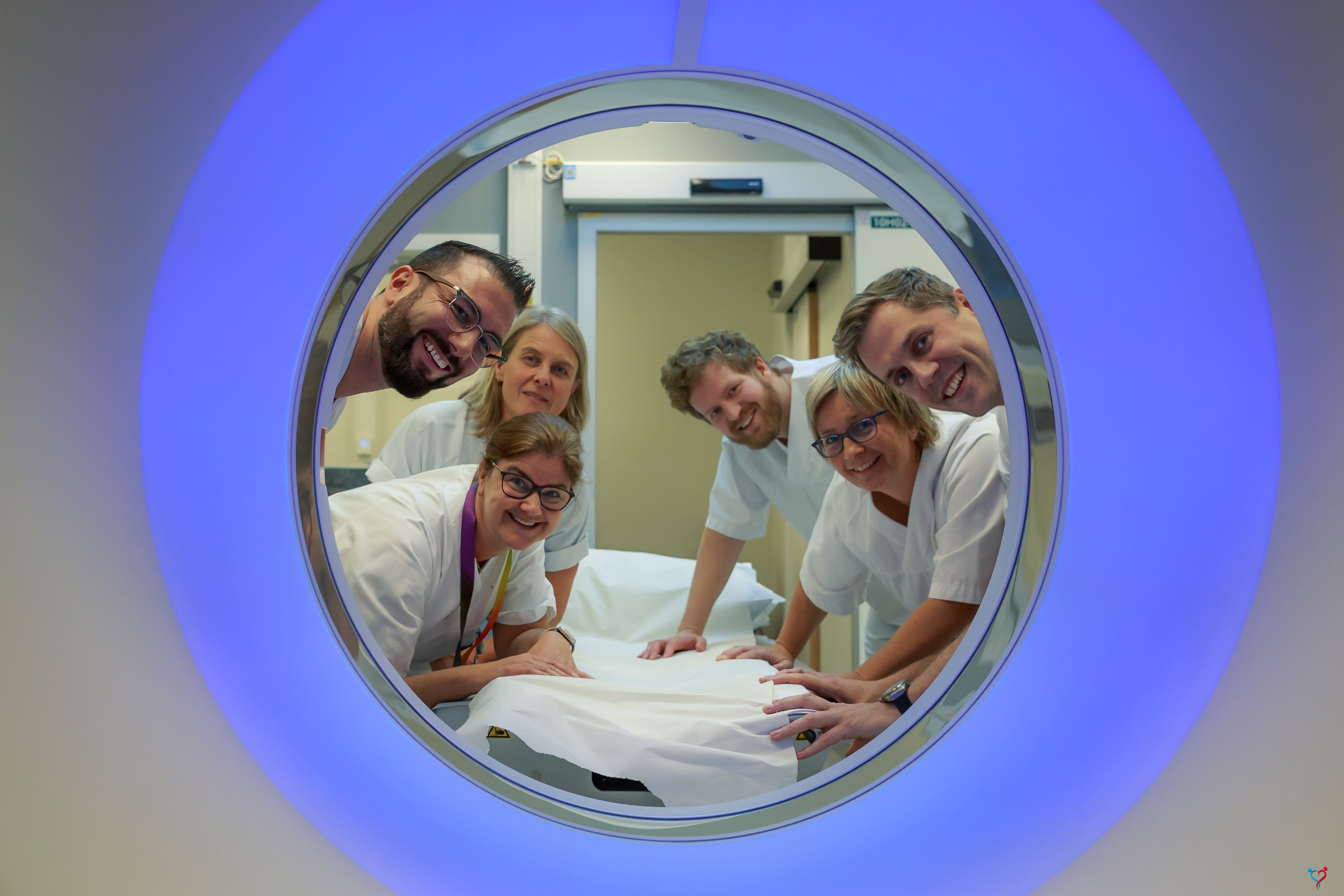 Seks smilende mennesker ikledd hvitt sykehustøy som ser gjennom en runding mot fotografen. Rundingen er en CT-maskin. Den avgir blått lys.