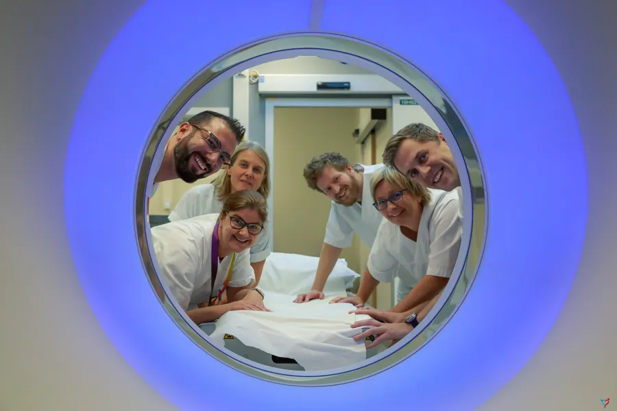 Seks smilende mennesker ikledd hvitt sykehustøy som ser gjennom en runding mot fotografen. Rundingen er en CT-maskin. Den avgir blått lys.