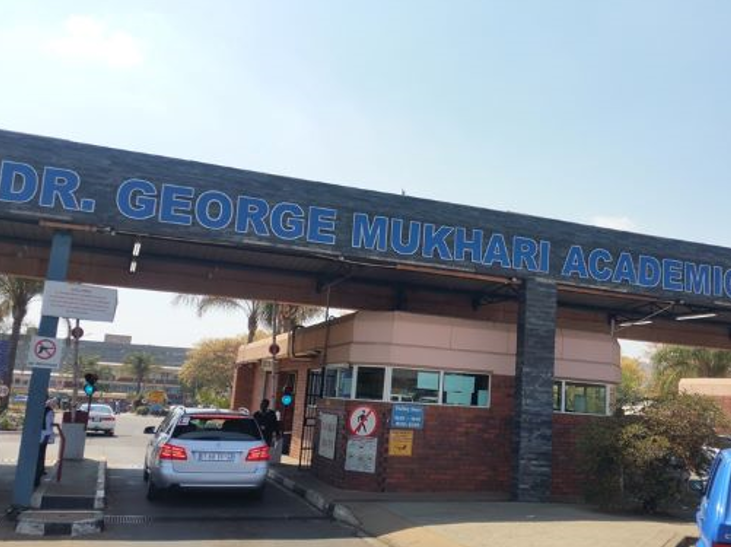 Foto. Innkøyrsel til sjukehusområde, svært navneskilt, forbodsskilt mot våpen og gåande, bilar. 