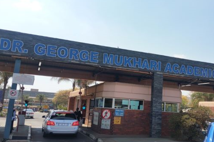 Foto. Innkøyrsel til sjukehusområde, svært navneskilt, forbodsskilt mot våpen og gåande, bilar.