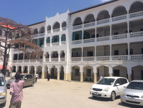 Eksteriør sykehusbygg Mnazi Mmoja Hospital i Zanzibar, parkeringsplass med tre, biler og folk. Foto