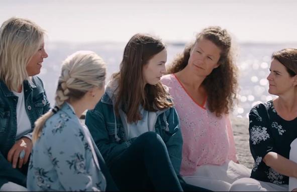 Ung jente sitter omringet av fire kvinner på et svaberg med havet i bakgrunnen. Foto