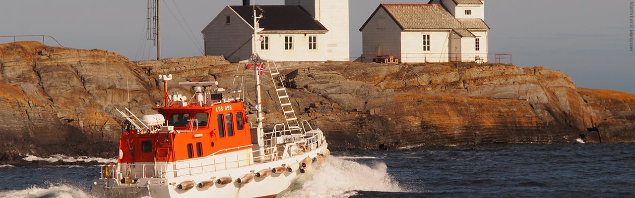 Foto. Bår på havet med fyrtårn i bakgrunnen.