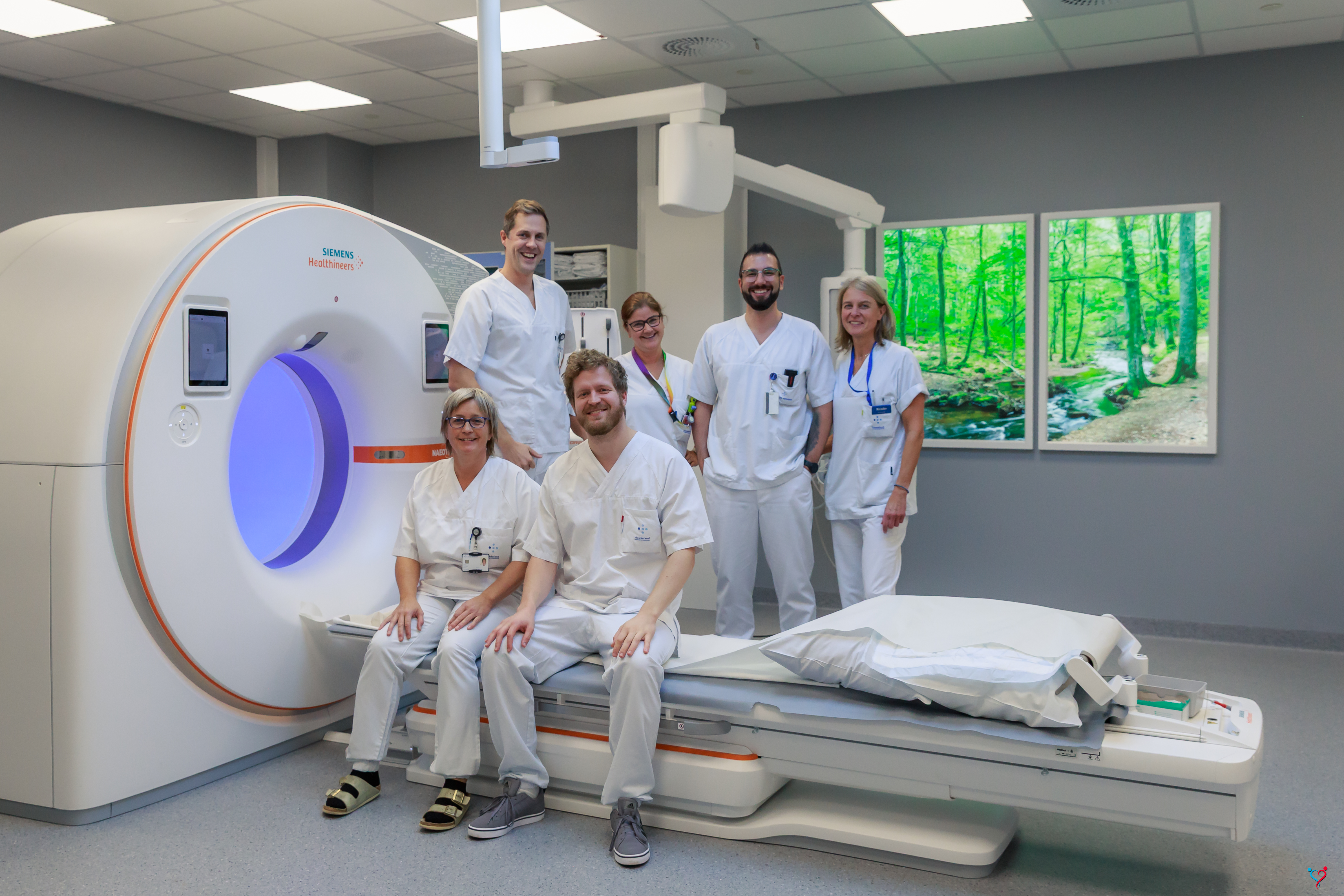 En CT-maskin der seks personer i sykehusklær poserer sammen sittende på CT-benken eller stående bak.