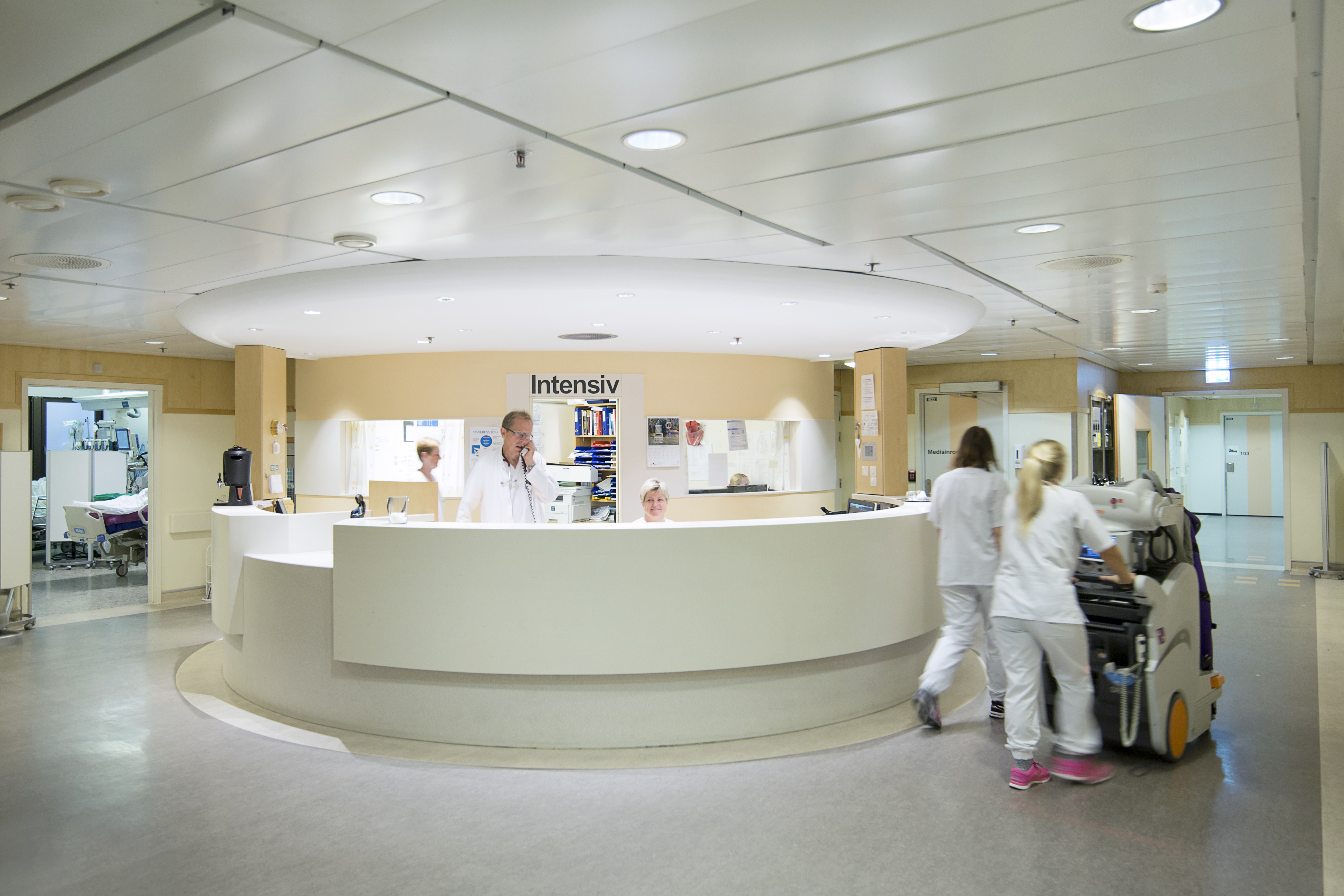 Viser en intensivsentral hvor en kvinne sitter å smiler bak skranken, mens en mann står og snakker i telefonen.