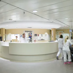 Viser en intensivsentral hvor en kvinne sitter å smiler bak skranken, mens en mann står og snakker i telefonen.