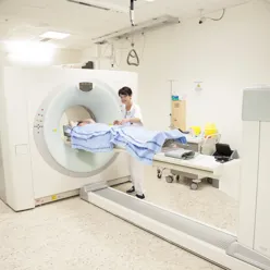 Kvinnelig helsepersonell gjør klar pasient som ligger på benk med teppe på seg for PET-undersøkelse. Foto