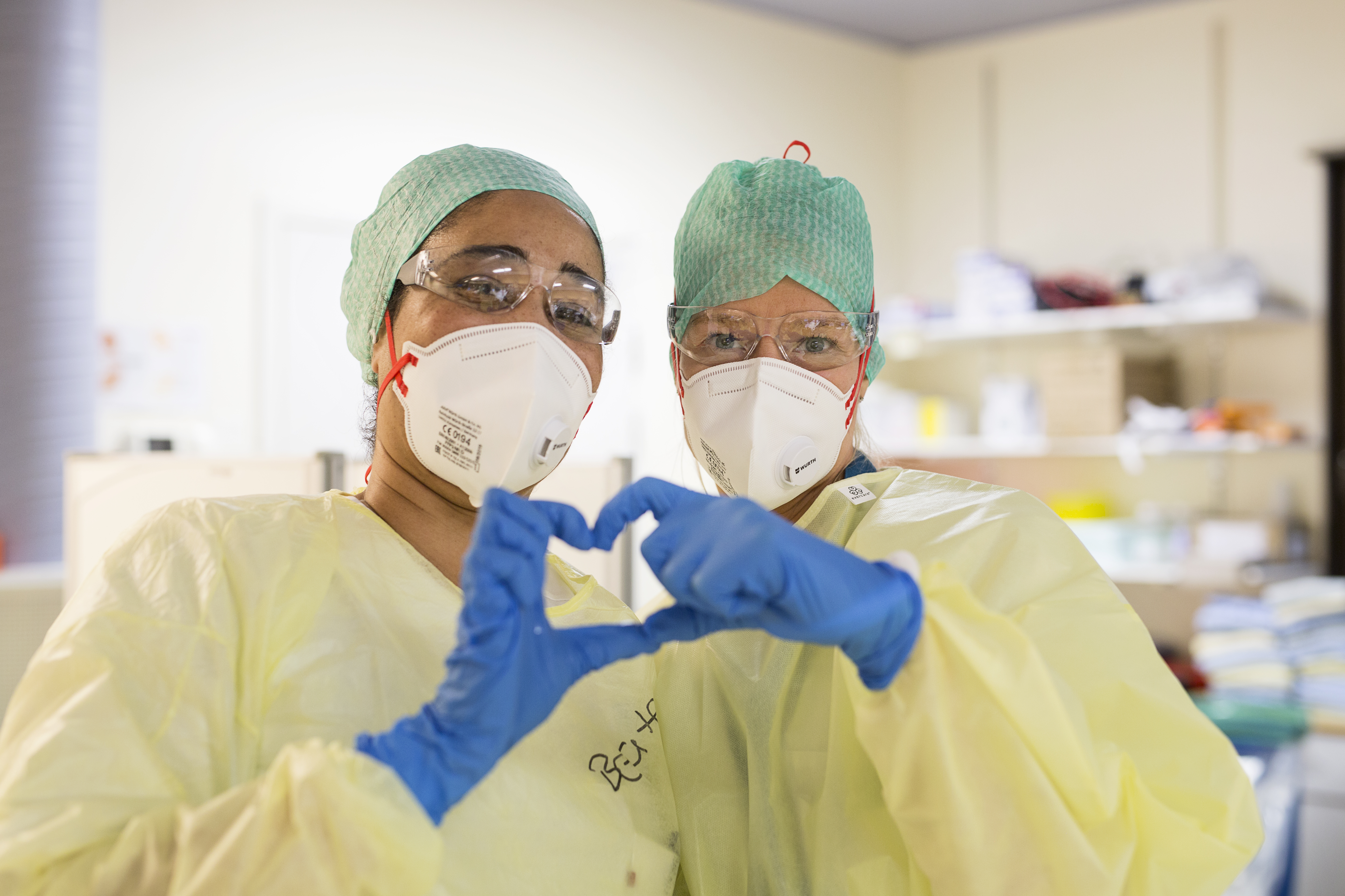 To helsepersonell ikledd smittevernutstyr former et hjerte sammen med hendene. Foto