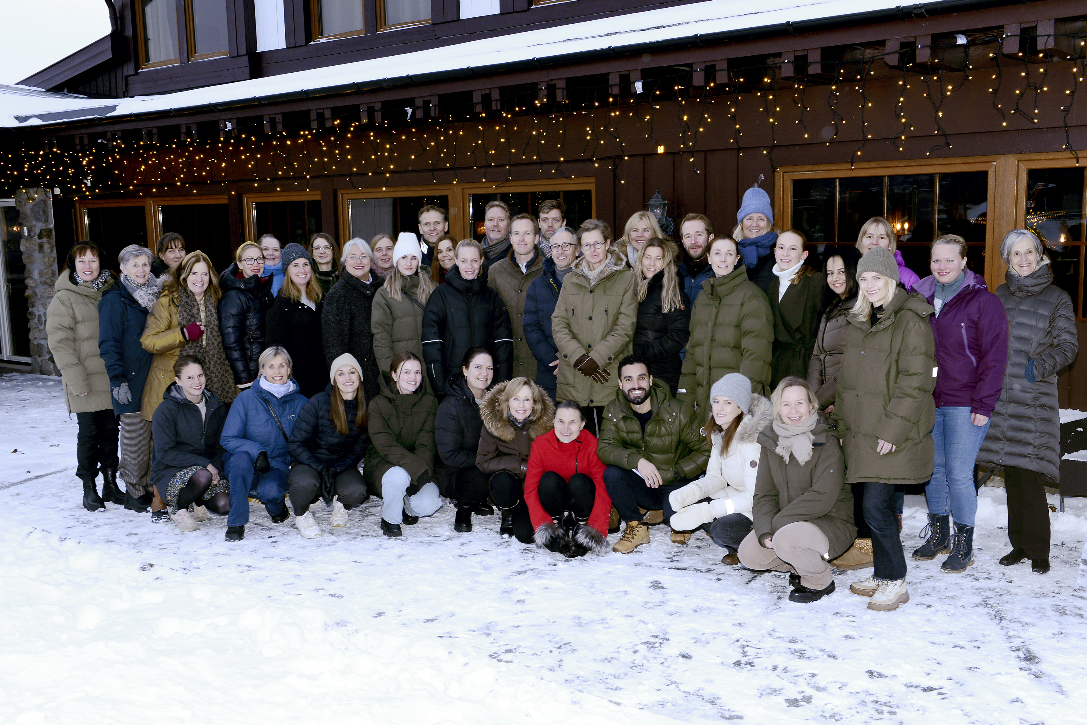 Gruppe mennesker kledd i vinterklær stilt opp for gruppebilde utenfor hus med julebelysning og snø på bakken. Foto