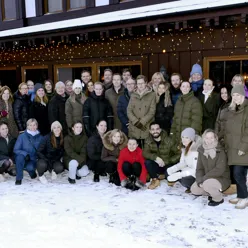 Gruppe mennesker kledd i vinterklær stilt opp for gruppebilde utenfor hus med julebelysning og snø på bakken. Foto
