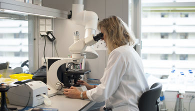 Kvinne i hvit frakk ser i mikroskop på laboratorium. Foto