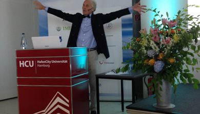 Foredragsholder på konferanse står foran rollup og slår ut med armene. Foto
