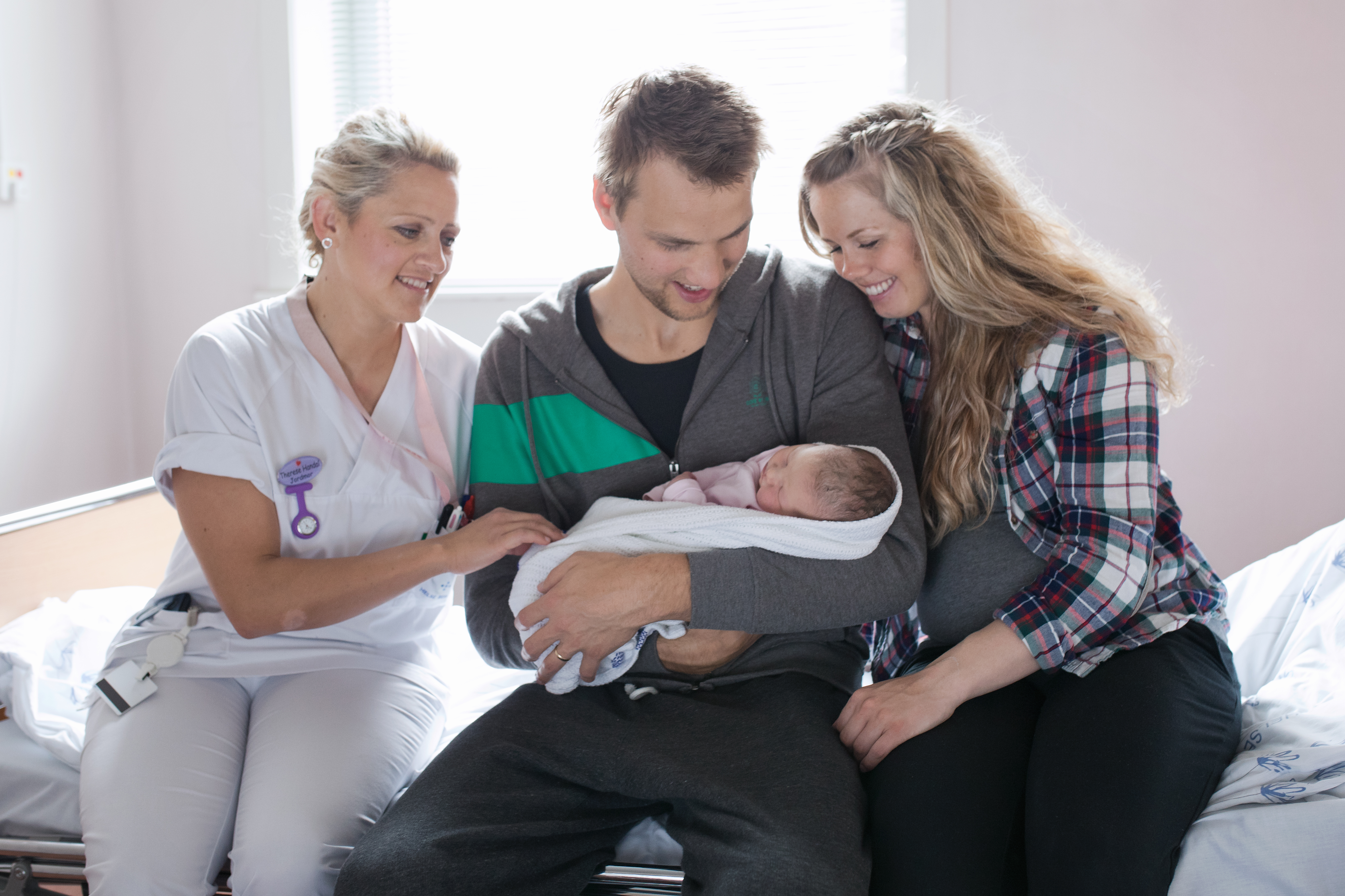 Jormor og nybakte foreldre sitter på seng, far med baby på armen. Foto