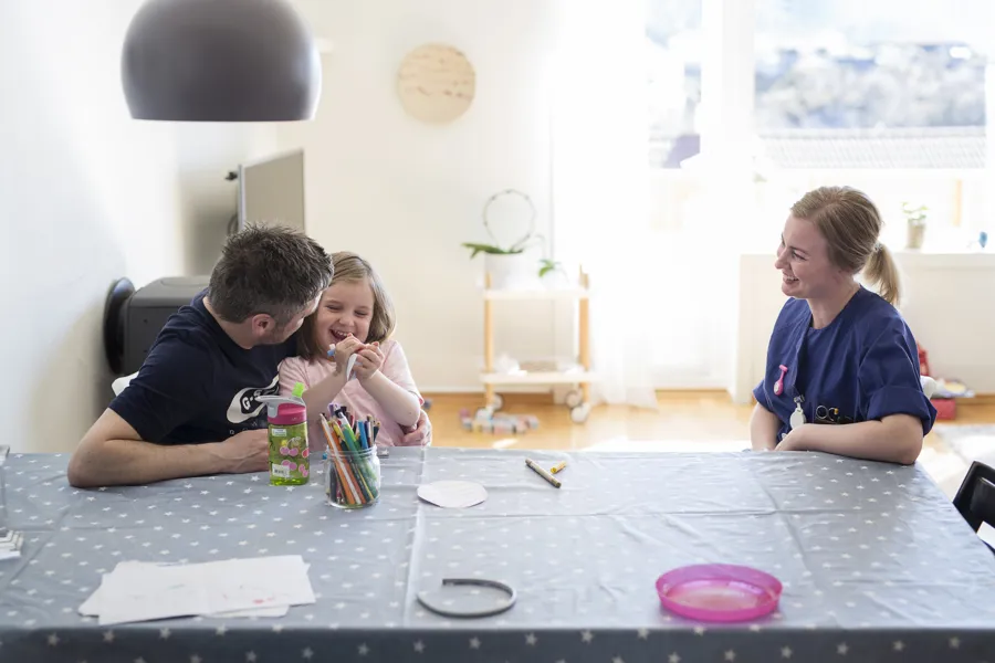 Far med datter på fanget sitter ved kjøkkenbord sammen med smilende sykepleier. Foto