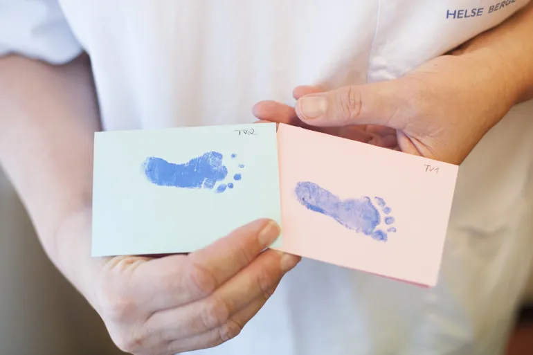 Nærbilde av helsepersonell sine hender som holder to ark med baby fotavtrykk. Foto