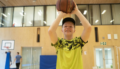 Ung gutt med gul tskjorte og caps med basketball i gymsal. Foto