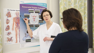 Kvinne i hvit uniform peker på plakat med anatomiske tegninger av nervesystem for kvinne som står med ryggen til. Foto