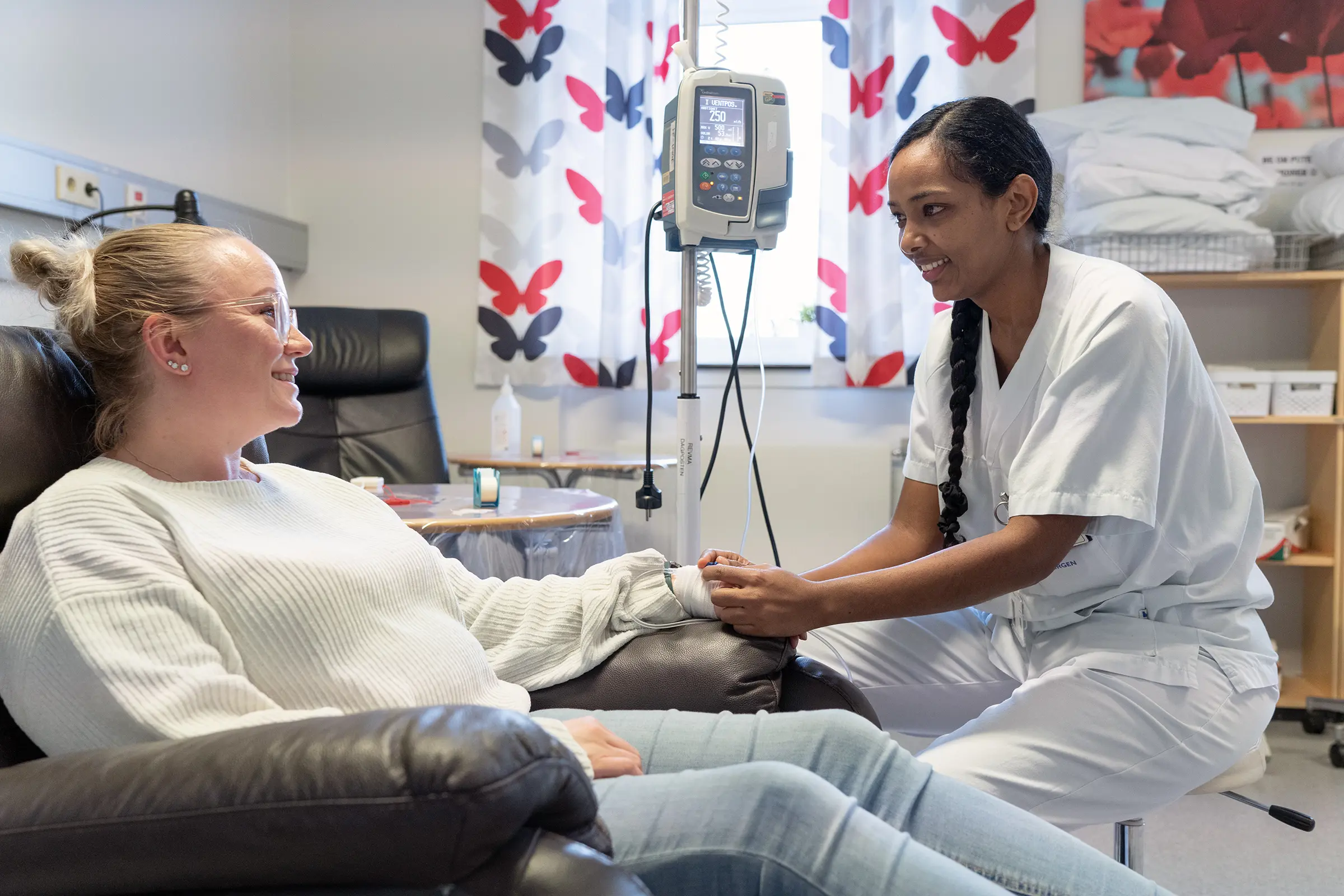 Pasient sitter i stol mens sykepleier gjør klar til pasient for infusjon. Foto