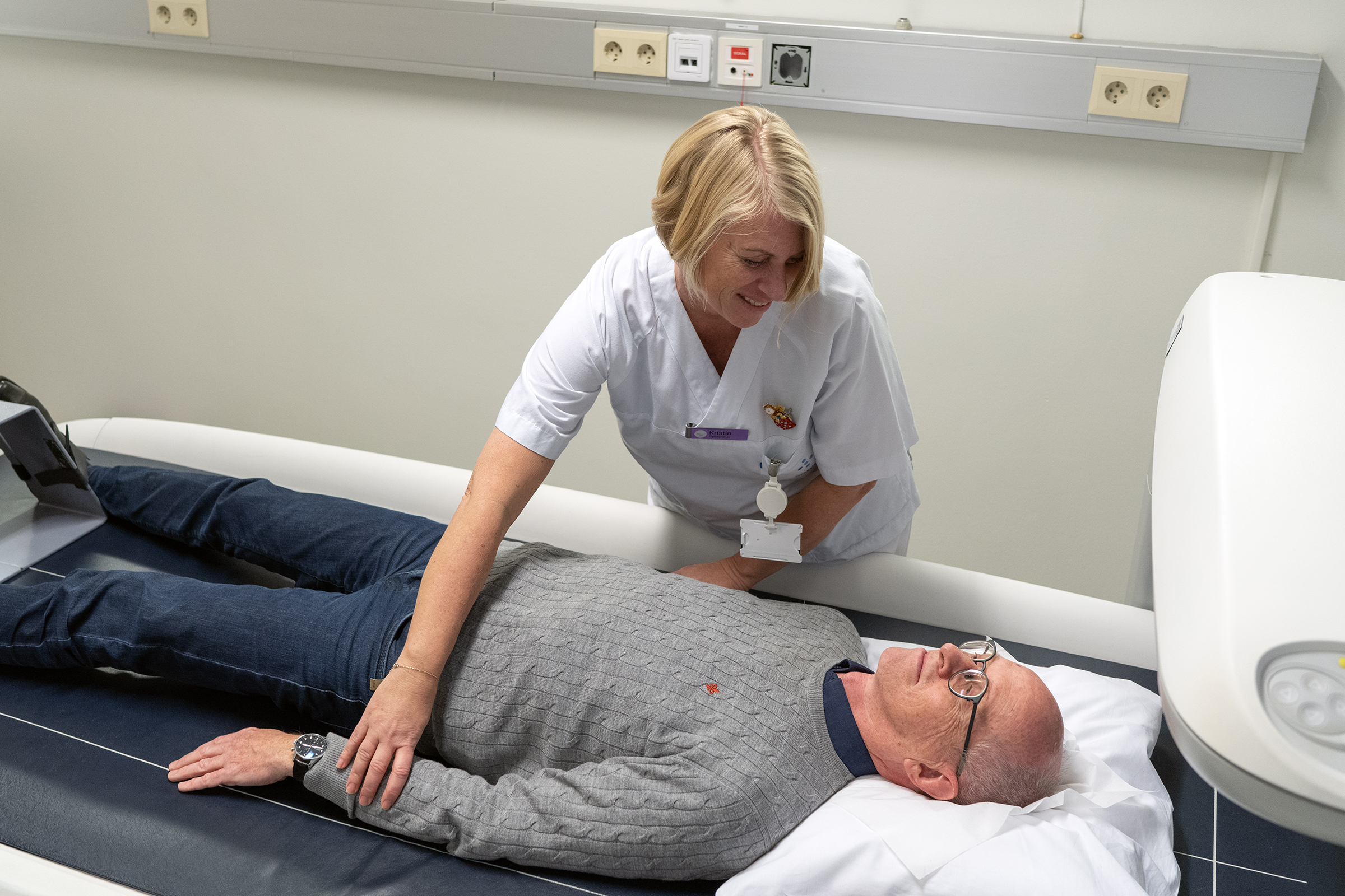 Kvinnelig helsepersonell støtter pasient som ligger på undersøkelsesbenk. Foto