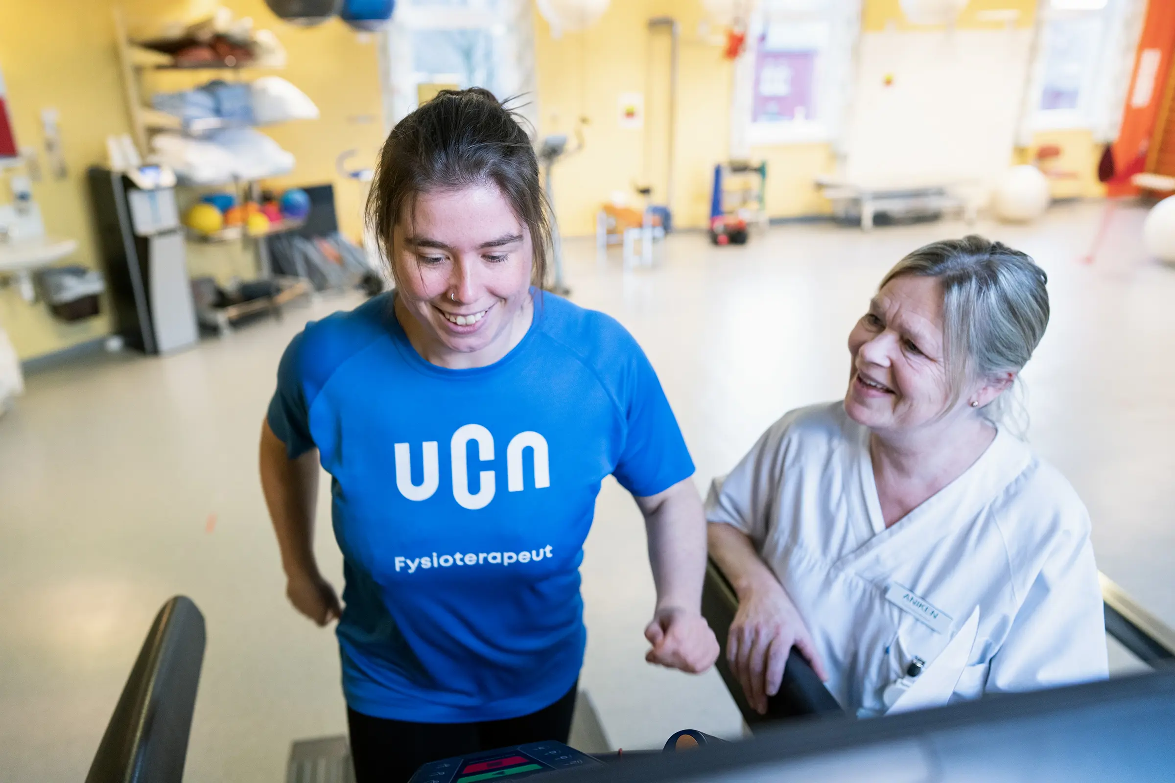 Blond fysioterapeut med hestehale, pasient med blå t-skjorte på tredemølle i treningsrom. Foto