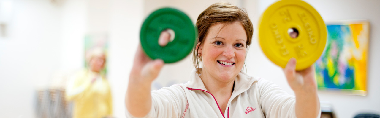 Smilende kvinne i treningsklær  holder opp ulike vekter mot kamera. Foto