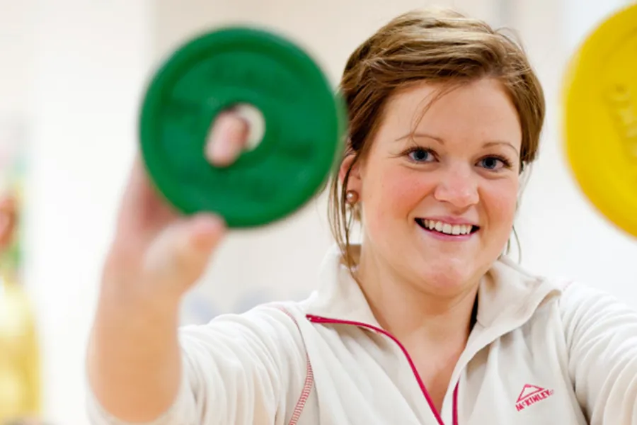 Smilende kvinne i treningsklær  holder opp ulike vekter mot kamera. Foto