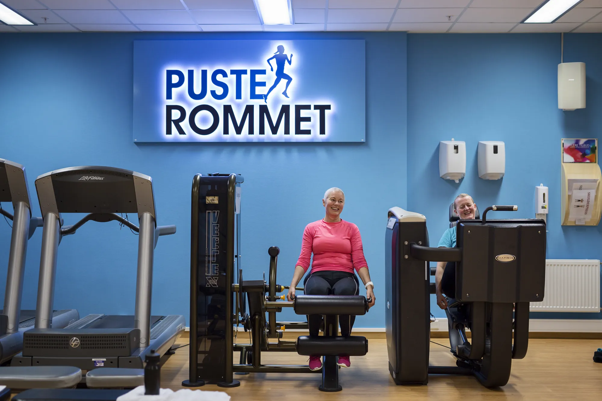 To damer i treningsklær sykler treningsrom med skiltet 