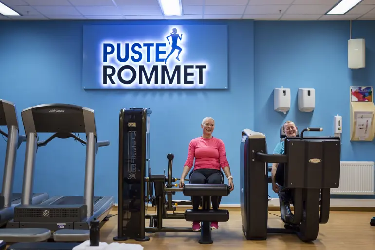To damer i treningsklær sykler treningsrom med skiltet