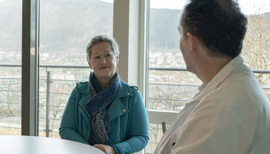 Eldre kvinne sitter i stol foran vindu, ser på mann i hvitt. Foto